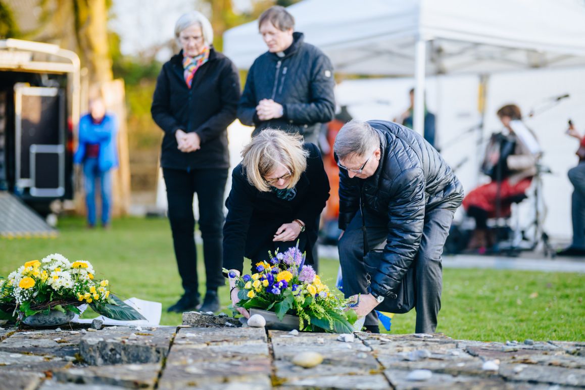 Anatoli Perov legt Blumen nieder, im Hintergrund die evangelische Pfarrerin und katholischer Pfarrer Wewelsburg 